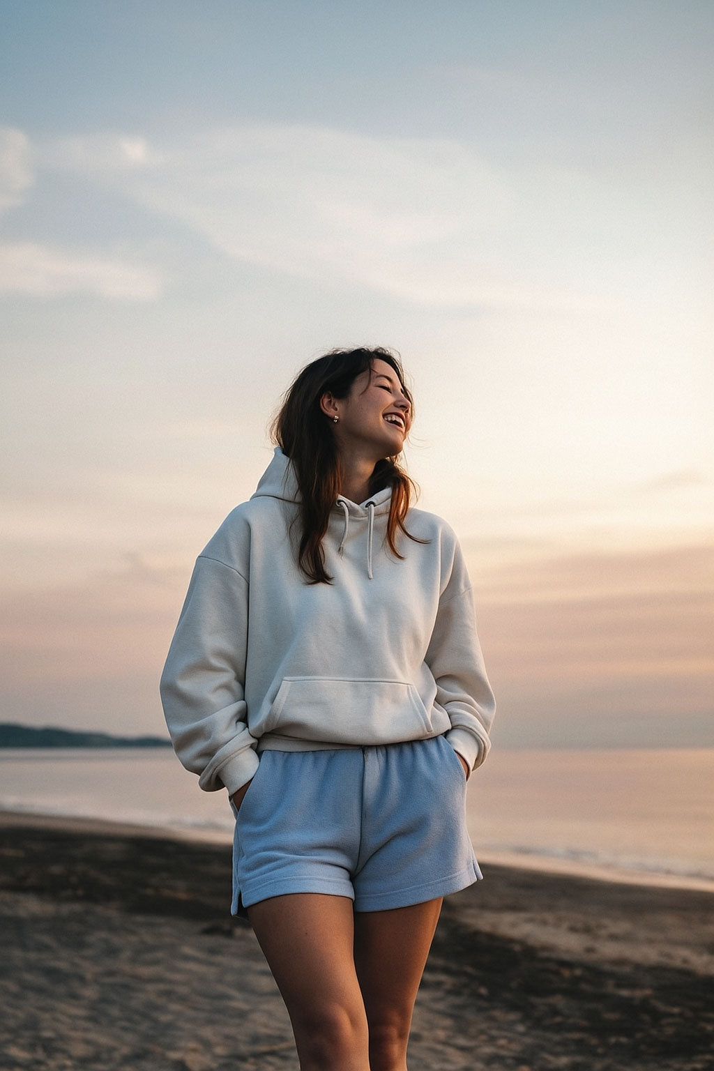 Woman in a light gray hoodie and shorts standing on a beach at sunset.